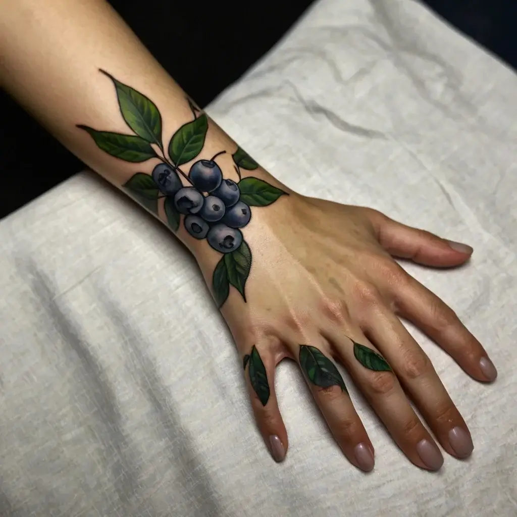 Tattoo of vibrant blueberries with green leaves on the wrist, extending to fingers with detailed leaf accents.