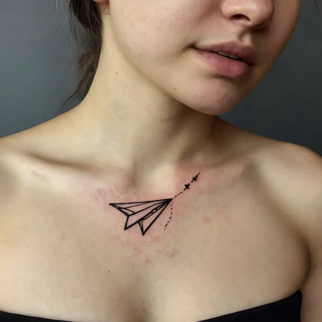 Tattoo of a minimalist paper plane with dashed trail on the collarbone, signifying freedom and adventure.