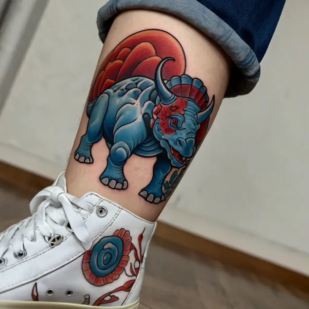 A vibrant tattoo of a blue triceratops with a red frilled crest, set against a deep red circular backdrop on a leg.