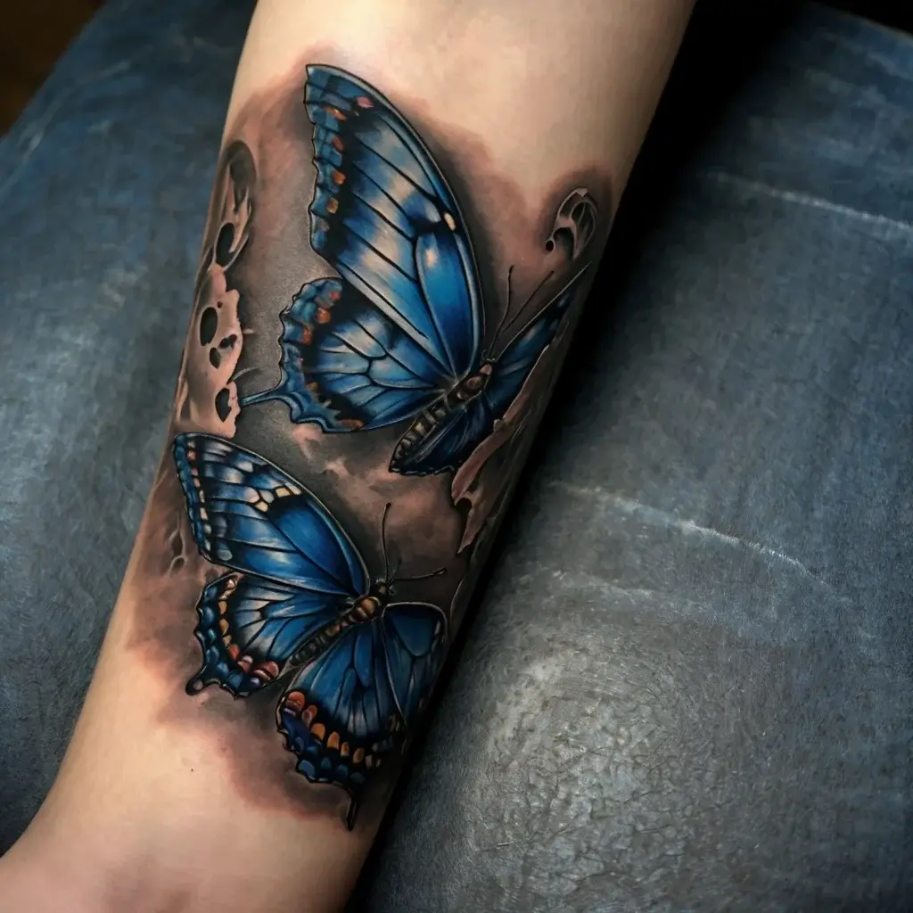 Tattoo of two vivid blue butterflies with detailed wings, surrounded by realistic shadows and floral elements on the forearm.